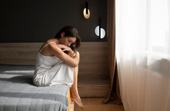 A young woman sitting on the edge of her bed in the morning, looking down with exhaustion and sadness, struggling to start the day.